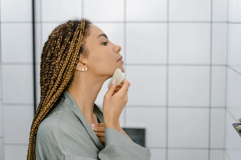 A woman using a gua sha stone along her jaw in a bathroom