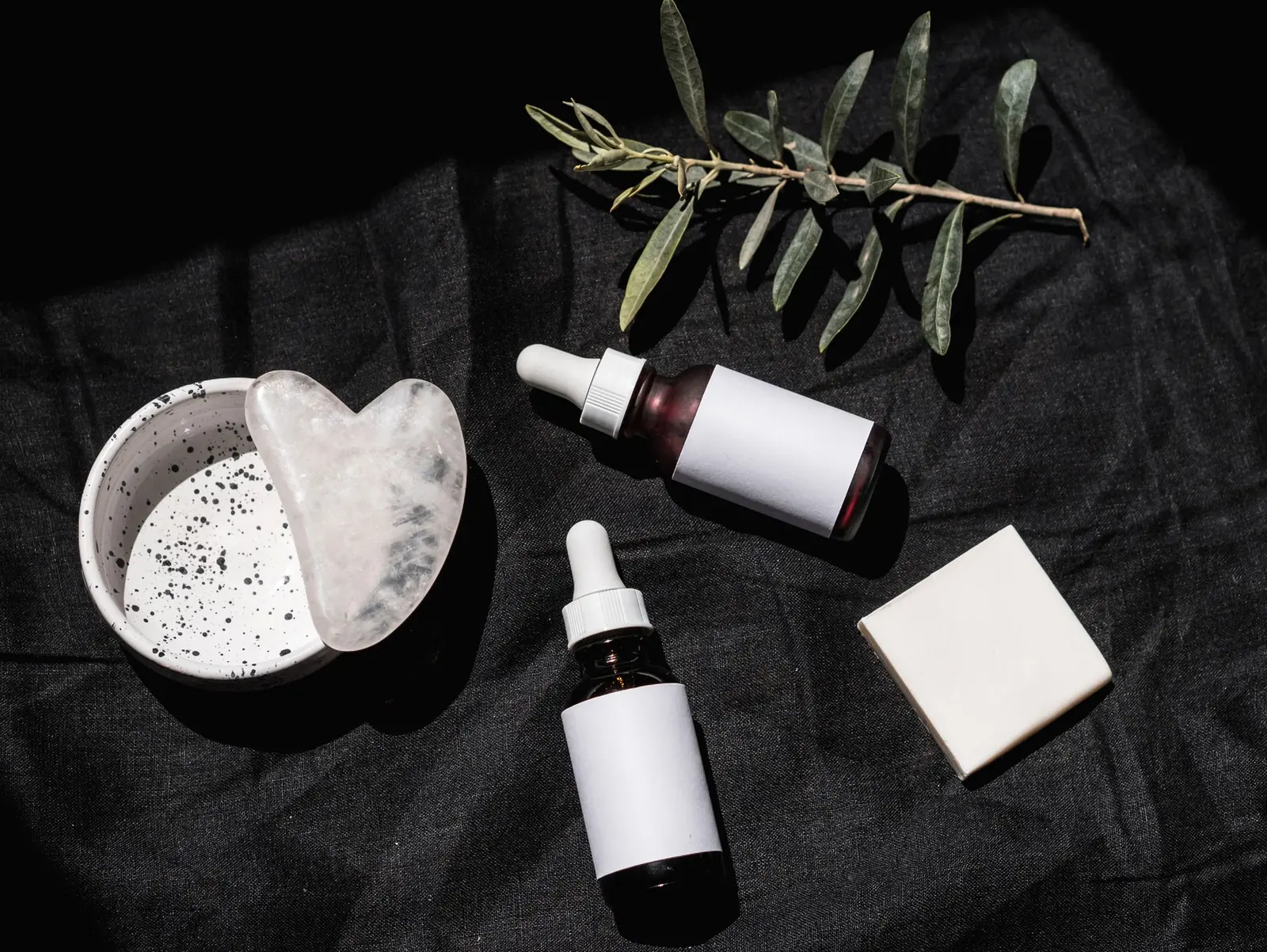 Brown glass skincare bottles, a gua sha stone and leaves on dark fabric