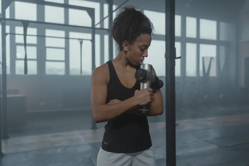 A woman using a massage gun on her upper arm in a gym