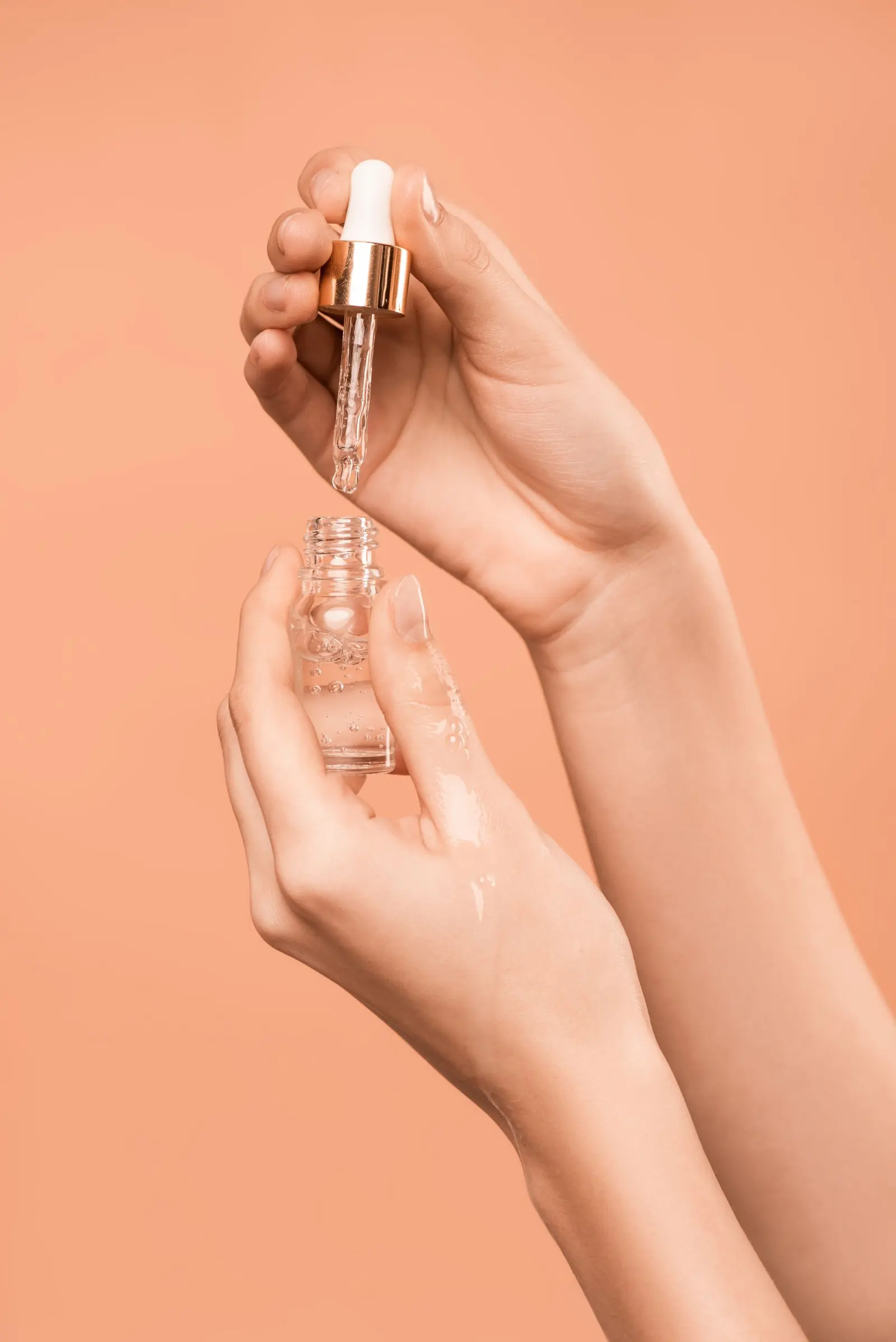 Hands holding a clear bottle and glass dropper against a peach background