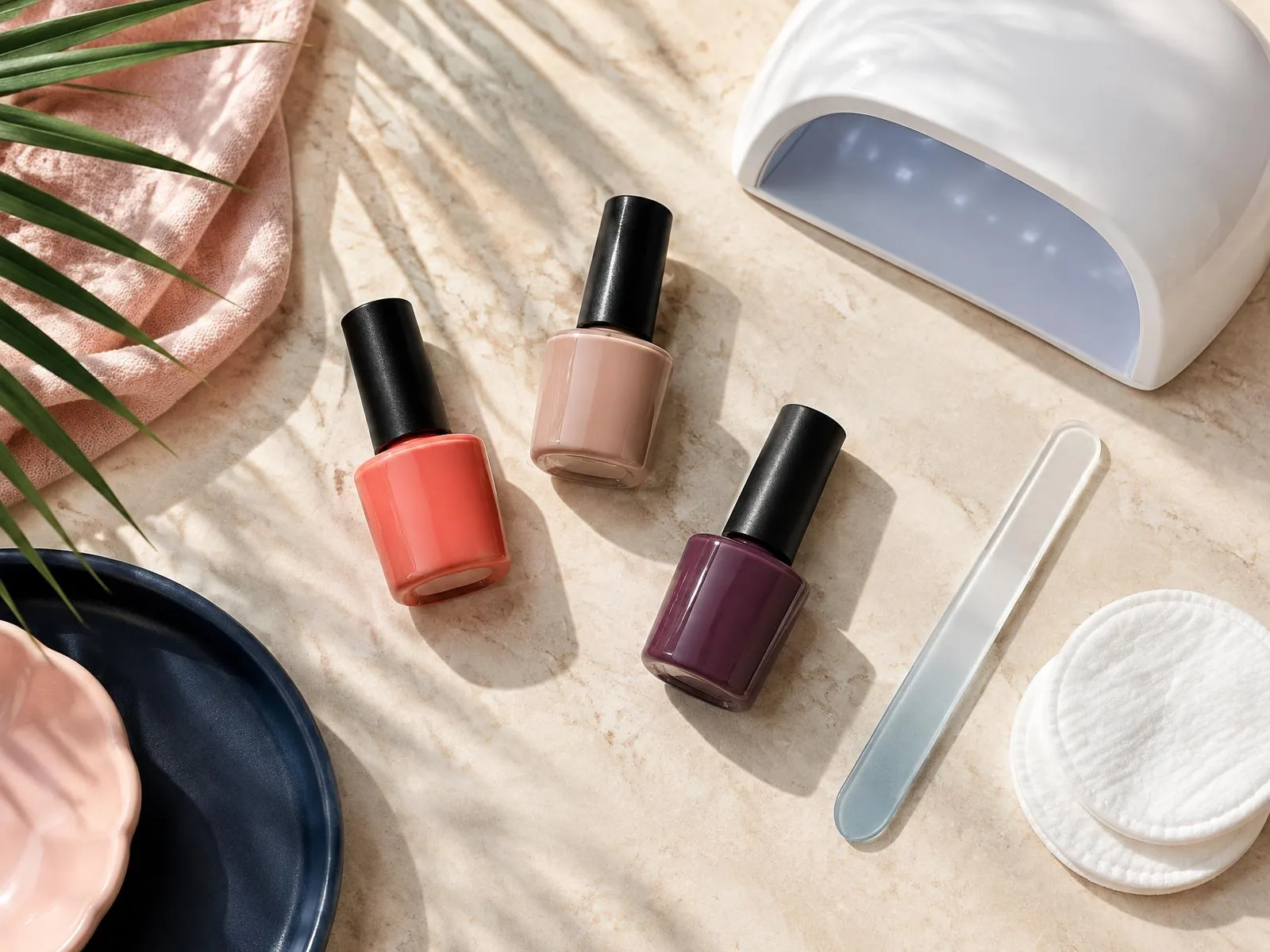 Unbranded coral, nude and plum gel polish bottles, a white nail lamp, a glass file and cotton pads on stone